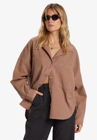 Bruine oversized shirt van zachte stof, met een button-down voorkant, twee borstzakken en lange mouwen. Zwarte broek maakt de look compleet.