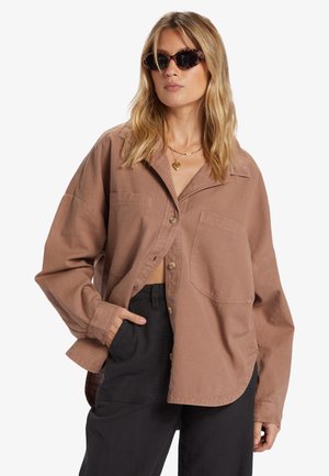 Hnědá oversized košile vyrobená z měkké tkaniny, s knoflíkovou přední částí, dvěma náprsními kapsami a dlouhými rukávy. Černé kalhoty doplňují celkový vzhled.