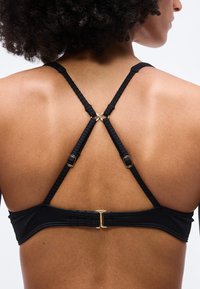 Πλάτη γυναίκας που φοράει μαύρο σουτιέν με ρυθμιζόμενους, σταυρωτούς ιμάντες και χρυσά μεταλλικά στοιχεία σε απλό λευκό φόντο.