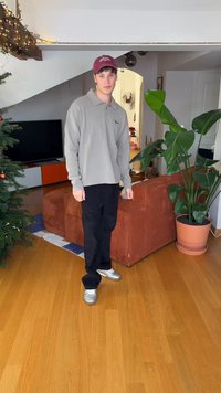 Grijze polo met lange mouwen, zwarte broek en zilveren sneakers. In een woonkamer met een plant, een bruine corduroybank en een tv. Vloer van licht hout.