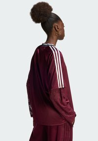 Αθλητικό τοπ βουργουνδί με μακριά, φαρδιά μανίκια, λευκές τρεις ρίγες στους ώμους και υφασμάτινες λεπτομέρειες κοντά στις μανσέτες.