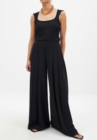 Zwarte mouwloze top met een vierkante halslijn, gecombineerd met zwarte wijde broek met plooien. Accentueert door een amberkleurige ketting.