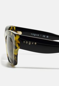 Fekete és teknős páncél mintás kerettel rendelkező napszemüveg, lapos formával és átmenetes lencsékkel. Arany "Vogue" logó az ágon. CE jelölés.