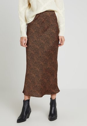 Femme portant une jupe midi à motifs noirs et marron, un pull en maille épaisse blanc et des bottines noires, debout sur un fond uni.