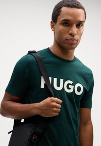 T-shirt de algodão verde com grande texto branco "HUGO". Modelo segura uma bolsa pequena de ombro preta com uma alça texturizada. Tecido suave, gola redonda.