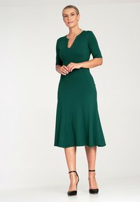 Sukienka do kolan w dopasowanym kroju, w kolorze zielonym, z dekoltem w kształcie litery V, krótkimi rękawami i rozkloszowaną spódnicą. Gładka tekstura bez widocznych wzorów. Czarne sandały na kostkę na obcasie.
