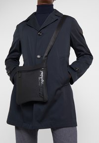 Černá crossbody taška s hladkým povrchem, s uzávěrem na zip a potiskem loga. Nesená přes tmavý kabát s límcem.
