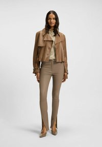 Braune Lederjacke im Cropped-Design mit doppelter Knopfleiste, kombiniert mit eng anliegenden beigefarbenen Hosen mit seitlichen Schlitzen und spitzen Absatzschuhen.