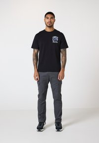 Czarny bawełniany t-shirt z grafiką na lewej piersi, zestawiony z szarymi spodniami cargo z bocznymi kieszeniami i elastycznymi mankietami. Czarne trampki.