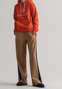 Sweat à capuche orange avec logo blanc, associé à un pantalon large couleur sable avec des bandes latérales bleu marine. Chaussures noires complètent la tenue.
