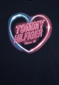 T-shirt en coton bleu marine avec un graphisme en forme de cœur néon, le texte "TOMMY HILFIGER" en lettres majuscules et le texte "Since 85", des accents vibrants en bleu et rose.