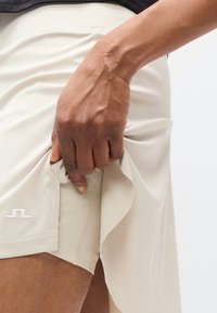 Αθλητική φούστα σε ανοιχτό μπεζ χρώμα με πλευρική χαραγή, που αναδεικνύει ένα απαλό, ελαφρύ ύφασμα. Λευκή λεπτομέρεια με λογότυπο στο κάτω αριστερό μέρος.