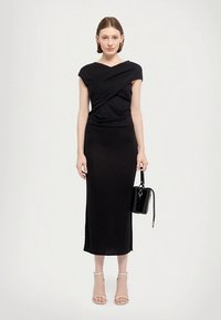 Rochie neagră, mulată, până la genunchi, cu mâneci scurte tip căpăcel și design drapat. Asortată cu o geantă mică neagră și tocuri transparente cu barete.