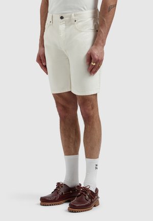Man wearing off-white denim shorts, white crew socks, brown leather boat shoes, and a white shirt, standing against a plain background.