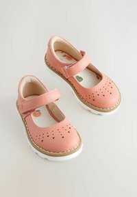 Różowe skórzane buty typu Mary Jane z rzepem, perforacjami w kształcie serca na czubku, białą podeszwą i wyściełanym wnętrzem.
