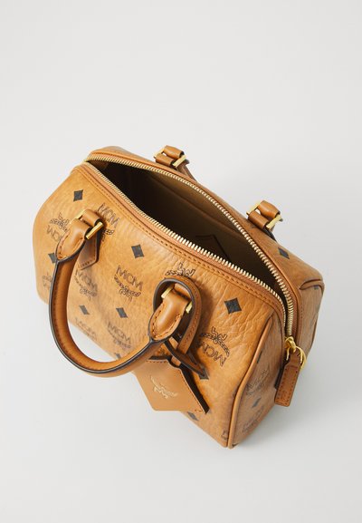 Tan læder MCM håndtaske med sorte logoer, guld lynlås delvist åben, rundede håndtag og en lædermærke på en hvid overflade.