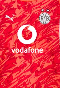 Punainen urheilusydäntoppi, joka esittää rohkeaa, abstraktia kuviota, sisältää Vodafonen logon ja Borussia Dortmundin vaakunan kahdella tähdellä. Valmistettu joustavasta kankaasta.