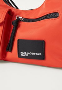 Crvena najlonska torba s crnim patentnim zatvaračem i crnom etiketom s logotipom "KARL LAGERFELD JEANS" u bijeloj boji. Tekstura s završnom obradom i izdržljiva oprema.