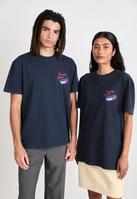 Camisetas de algodón azul marino con manga corta, que presentan un logo gráfico en rojo y azul en el pecho. Corte relajado, llevadas por dos modelos.