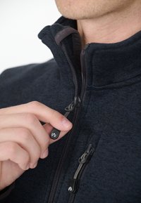 Tummansininen vetoketjullinen fleece-takki, jossa on teksturoitu kangas, korkea kaulus ja koristeelliset vetoketjun vedinlogot. Käsi puristaa vetoketjua.