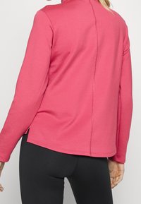 Rosa långärmad topp med hög krage, med rak fåll, sidospalter och synlig sömnadsdetalj längs ryggen.