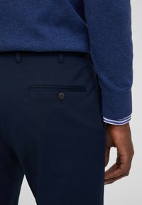 Pantalones azul marino con un bolsillo trasero que cuenta con un cierre de botón. El conjunto se complementa con un suéter azul suave con tejido texturizado.