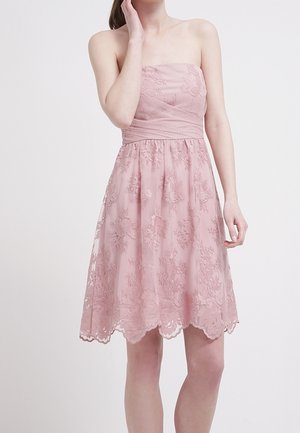 Robe rose sans bretelles longueur genou avec superposition en dentelle florale et taille cintrée, portée par une femme debout sur un fond uni.