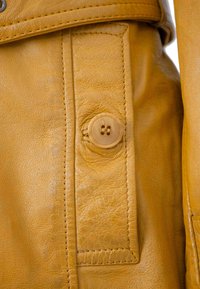 Gelber Lederjackendetail mit großem Knopf, sichtbarer Naht und strukturierter Oberfläche. Der Knopf ist hellfarben und hat zwei Löcher.