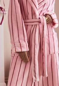 Pink fleece robe featuring vertical red stripes, a shawl collar, wrap design, and tied belt. Includes front pockets with a soft texture.