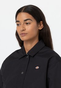 Giacca nera con bottoni e colletto, caratterizzata da un piccolo logo Dickies sulla parte sinistra del petto e una texture liscia.