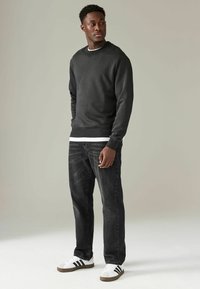 Sudadera negra con puños de canalé, llevada sobre una camisa blanca; combinada con jeans oscuros y zapatillas blancas con rayas negras.