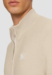 Beige Zip-Up-Pullover mit hohem Kragen, gerippter Textur und weißem Logo-Patch. Sichtbarer Reißverschlussdetails entlang der Vorderseite. Nahansicht.