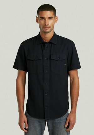 Uomo con capelli corti che indossa una camicia nera a maniche corte con due tasche sul petto e jeans blu su sfondo semplice.