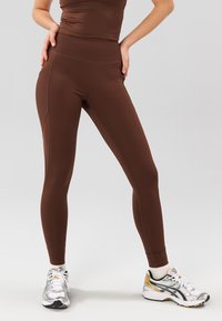 Leggings atléticos marrones con cintura alta, que presentan una textura suave y un diseño ajustado. Combinados con zapatillas de correr plateadas.