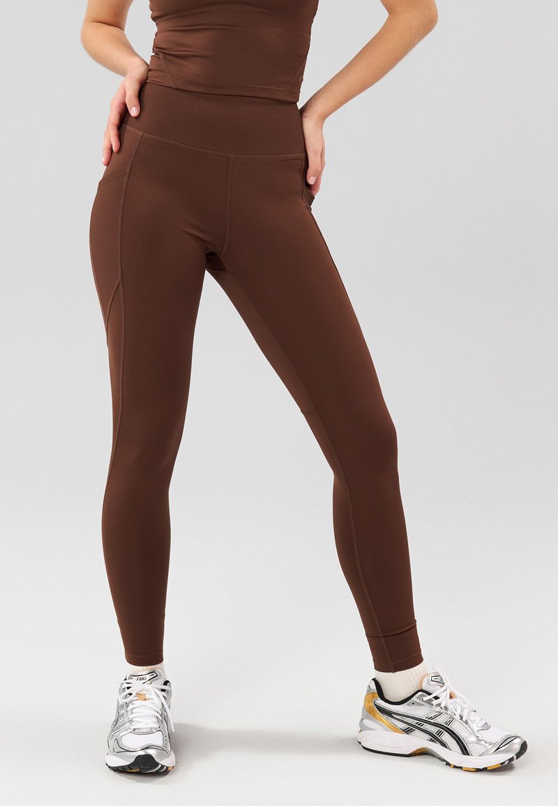Leggings atléticos marrones con cintura alta, que presentan una textura suave y un diseño ajustado. Combinados con zapatillas de correr plateadas.