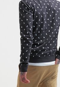 Sudadera negra con patrones geométricos y temáticos de la naturaleza en blanco, con cuello y puños acanalados, y un corte relajado.