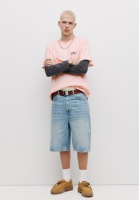 Tricou roz deschis cu un design grafic, suprapus peste o cămașă lungă închisă. Șorturi din denim albastru și pantofi maro cu șosete albe completează lookul.
