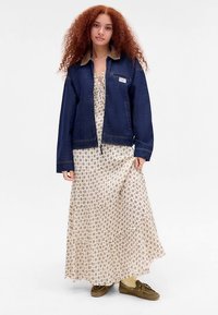 Jeans jakna z mehkim ovratnikom, združena z dolgo obleko s kremno-rjavim vzorcem in rjavimi čipkastimi čevlji.