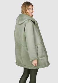 Eine lächelnde Frau mit welligem blonden Haar trägt einen übergroßen, hellgrünen Lammfellmantel mit Kapuze und seitlichen Reißverschlusstaschen, kombiniert mit dunklen Leggings.