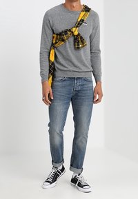 Homme portant un pull gris, un jean bleu retroussé, des baskets noires et blanches, avec une chemise à carreaux jaune et noire nouée sur la poitrine.