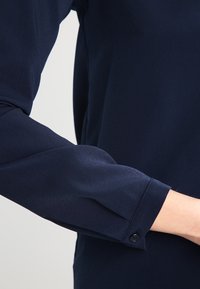 Mörkblå blus med en slät textur, som har en knappad manschett och en skräddarsydd passform. Ärmen är något samlad vid axeln.