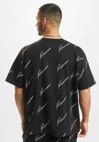 Juodas marškinėliai su pakartotiniu baltu „Rocawear“ logotipu kreivai šriftu. Trumpomis rankovėmis ir laisvu siluetu. Lygus audinio tekstūros pojūtis.