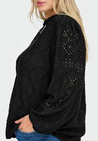 Blusa nera in pizzo con motivi floreali testurizzati, maniche lunghe a sbuffo e scollatura arricciata. Il tessuto è leggero e semitrasparente.