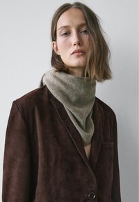 Szalik z khaki wełny elegancko opada na ciemnobrązową zamszową marynarkę, charakteryzującą się gładką fakturą i klasycznym klapem z pojedynczym guzikiem.