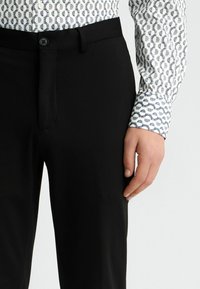 Schwarze Hose mit schmaler Passform, mit Knopfverschluss und Gürtelschlaufen, kombiniert mit einem weißen Hemd mit einem kreisförmigen blauen Muster.