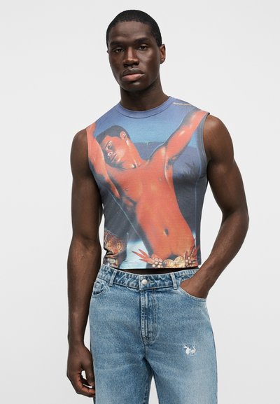 Αμάνικο γραφικό μπλουζάκι με καλλιτεχνικό σχέδιο κορμού σε μπλε φόντο, συνδυασμένο με ανοιχτόχρωμο τζιν παντελόνι με λεπτομέρεια λογότυπου.