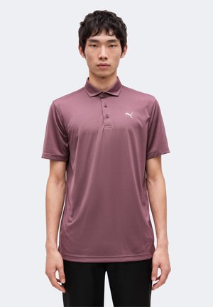 Mann trägt ein mauvefarbenes kurzärmeliges Poloshirt mit kleinem weißem Puma-Logo auf der linken Brust und steht vor einem einfarbigen hellen Hintergrund.