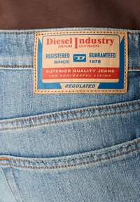 Šviesiai mėlynos džinsinės kelnės turi odinį etiketę su raudonais ir mėlynais įrašais, nurodančiais "Diesel Industry", "Aukštos kokybės kelnės" ir registracijos duomenis.
