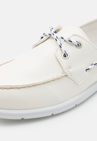 Weißes Lederbootsschuh mit glatter Oberfläche, ausgestattet mit blau-weißen geflochtenen Schnürsenkeln, Metallösen und einer weißen Gummisohle.