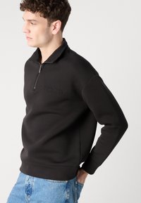 Hanorac negru cu fermoar, realizat dintr-un material moale, având un guler înalt și logo-ul „Calvin Klein” embosat pe piept, purtat cu blugi de culoare albastră deschisă.